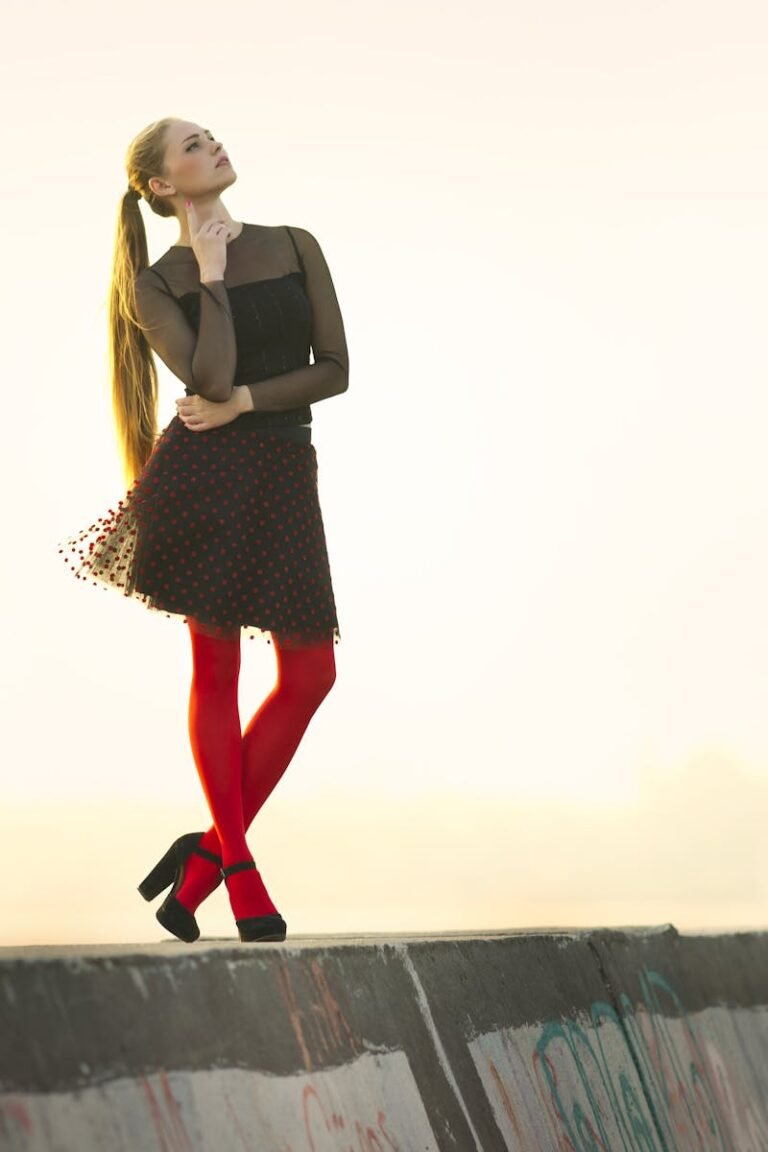 A fashionable woman in a black dress and red tights poses on a concrete barrier outdoors during the day.