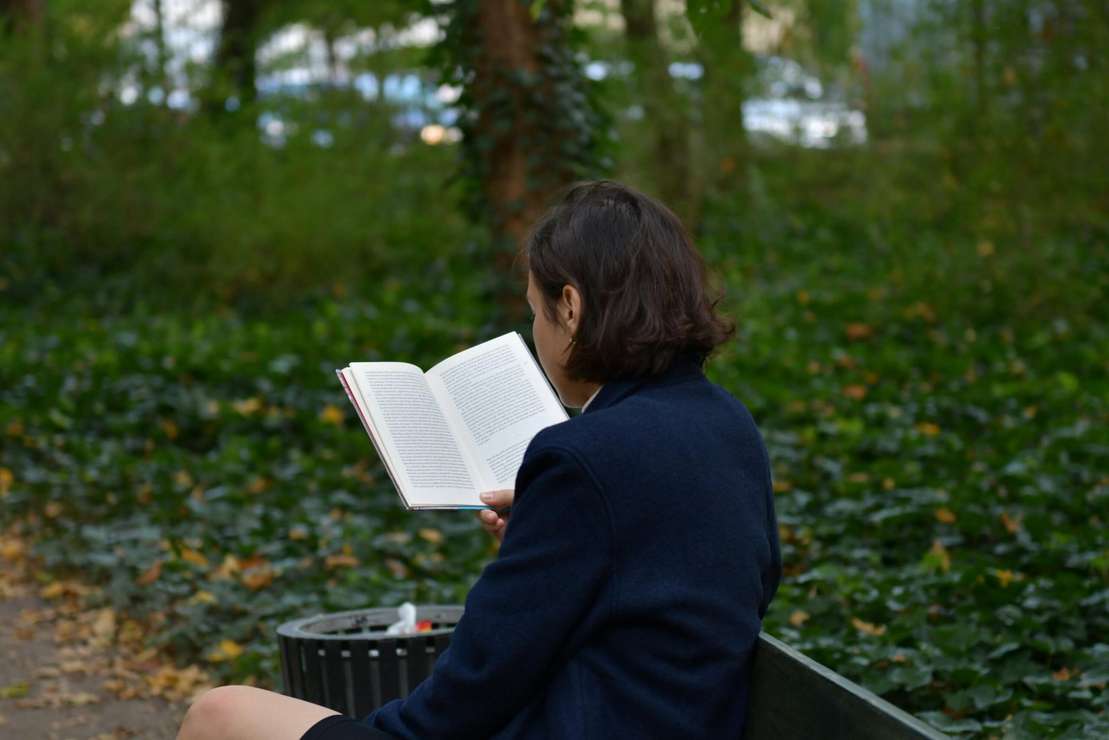 Back view of woman reading book outdoors in lush park setting.